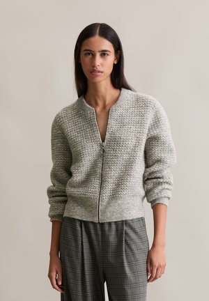 Cardigan grigio chiaro con zip, caratterizzato da un motivo a zigzag testurizzato, maniche e orlo a costine, abbinato a pantaloni a quadri grigi.