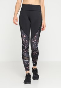 Černé legíny s vysokým pasem a růžovými květinovými a ptáčími vzory na dolních nohách, spárované s černými teniskami Nike.