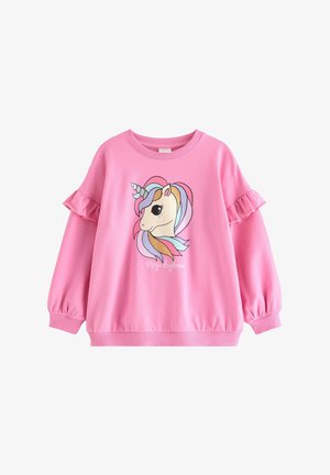 Jeune fille portant un sweat-shirt rose avec un imprimé licorne et un legging noir avec des motifs assortis de licorne et de cœur, posant sur un fond uni.