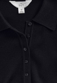 Zwarte geribbelde gebreide poloshirt met knopen en een wit label met de tekst "THE SET M Made in Bangladesh" bij de kraag.