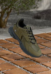 Olive green sneaker with mesh and suede materials, rounded shape, black laces, and a thick gray sole, displayed on a textured surface.