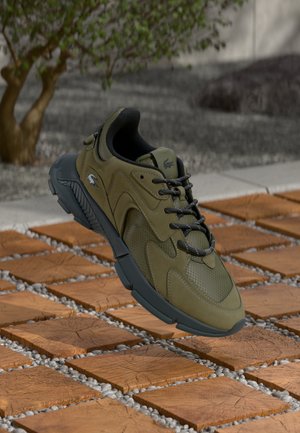 Olive green sneaker with mesh and suede materials, rounded shape, black laces, and a thick gray sole, displayed on a textured surface.
