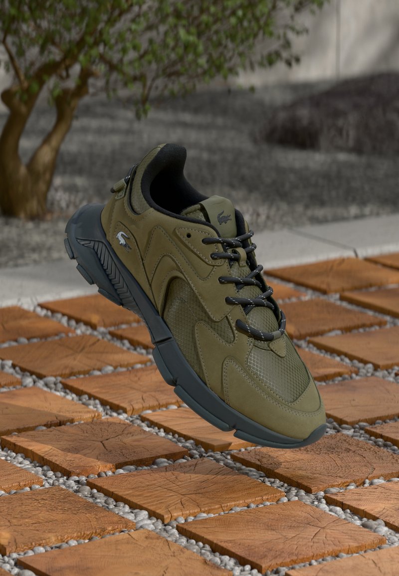 Olive green sneaker with mesh and suede materials, rounded shape, black laces, and a thick gray sole, displayed on a textured surface.