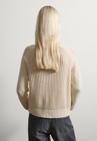 Suéter beige de punto con textura acanalada, cuello redondo y mangas largas. Presenta una cintura ajustada y costuras detalladas en los hombros.
