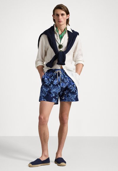Shorts de baie în nuanțe de albastru înflorate, cu șnur alb, asortați cu o cămașă din in albă și un pulover tricotat albastru așezat pe umeri. Eșarfă cu model verde.