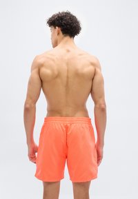 Νεαρός άνδρας με σγουρά μαλλιά, γυμνός από τη μέση και πίσω, φ οράει φωτεινά πορτοκαλί μαγιό, σε λευκό φόντο.