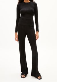 Černý žebrovaný top s dlouhým rukávem v kombinaci s úzkými černými kalhotami s bočními rozparky a černými sandály se čtvercovou špičkou na bosých nohou.