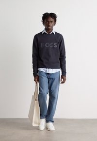 Σκούρο ναυτικό φούτερ με το λογότυπο "BOSS", λευκό πουκάμισο από κάτω, ανοιχτό γαλάζιο τζιν, λευκά αθλητικά παπούτσια και κρεμ τσάντα. Μινιμαλιστικός σχεδιασμός.