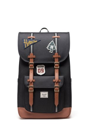 Sac à dos noir avec des sangles marron, des écussons décoratifs et une poche avant, posé debout sur fond blanc. Aucune personne présente.