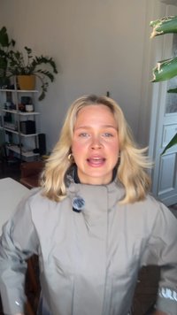 Blonde vrouw in een beige jas die binnenshuis spreekt, met planten en planken op de achtergrond.