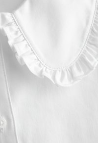 Cămașă albă cu guler volănat, textură fină și închidere cu nasturi pe față, punând accent pe un design curat și minimalist.