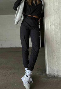 Person, der bærer sorte cargobukser, hvide sneakers, hvide sokker, sort kort top og grå jakke, stående i et betonet indendørs område.