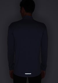Långärmad atletisk tröja i mörkblått tyg med hög krage, prydd med reflekterande detaljer vid bakre fållen för ökad synlighet.