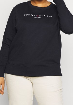 Czarna bluza Tommy Hilfiger z długim rękawem z logo i napisem "EST. 1985", noszona z jasnymi spodniami, osoba przycięta na wysokości szyi i kolan.