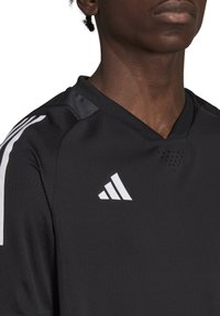 Zwart sportshirt met V-halsontwerp, voorzien van witte drie-strepen accenten en logo. Materiaal heeft een gestructureerde afwerking met ventilatiegaten.