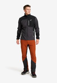 Man i svart och grå jacka samt rostorange utomhusbyxor, stående och tittande åt sidan, mot vit bakgrund.