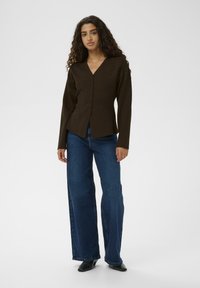 Brūns garās piedurknes kardigāns ar V veida kakla izgriezumu un priekšējo pogu aizdari, apvienots ar platām zilām džinsa biksēm un melniem apaviem.