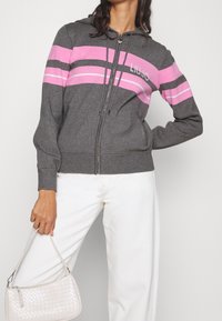 Szary rozpinany hoodie w różowe i białe poziome paski, z kapturem ze sznurkiem i przednimi kieszeniami. Modelka trzyma jasnoszary torebkę.