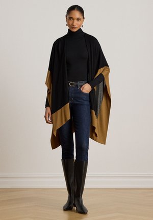 Femme portant un poncho bicolore noir et marron, un col roulé noir, un jean foncé et des bottes hautes noires, debout à l'intérieur.