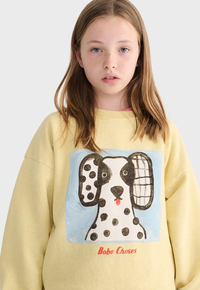 Dievča s svetlohnedými vlasmi nosí žltú mikinu s hravou ilustráciou škvrnitého psa a textom "Bobo Choses" pod obrázkom.