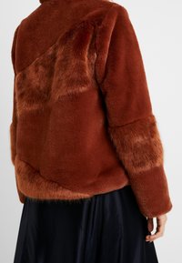 Femme portant une veste en fausse fourrure couleur rouille avec des textures de fourrure mélangées et une jupe noire, vue de dos à hauteur de la taille.