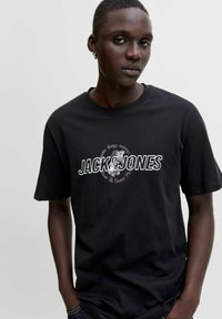 Noor mees tumeda nahaga, kandes musta "JACK & JONES" t-särki ja kõrvarõngaid, seisab lihtsa heleda tausta ees.