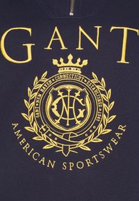Tissu marine avec écusson jaune brodé, couronne, couronne de laurier et texte mentionnant "GANT New Haven Connecticut Fondée en 1949 Vêtements de sport américains."