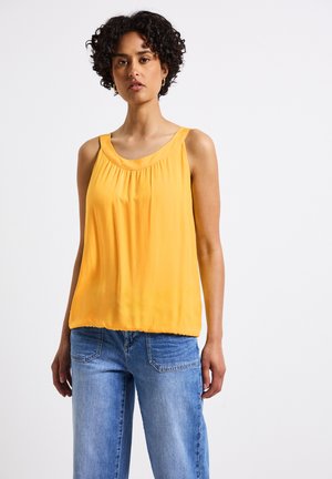 Top sans manches jaune avec un col rond, texture douce et détail froncé sous le col, associé à un jean bleu clair ample.