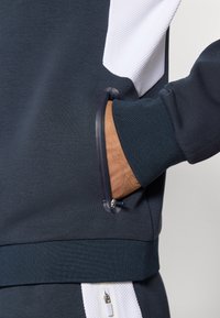 Tmavomodrá mikina s textúrovanou bielou časťou, zipsovou kapsou na boku a pružnými manžetami a lemom pre priliehajúci vzhľad.