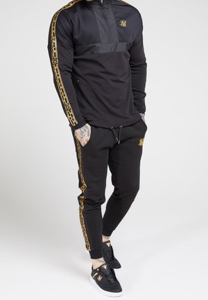 Mann in schwarzem Trainingsanzug mit goldenen Kettenmustern an den Ärmeln und an den Seiten der Hose, trägt schwarze Sneakers mit goldenen Akzenten und hat die Hände in den Taschen.