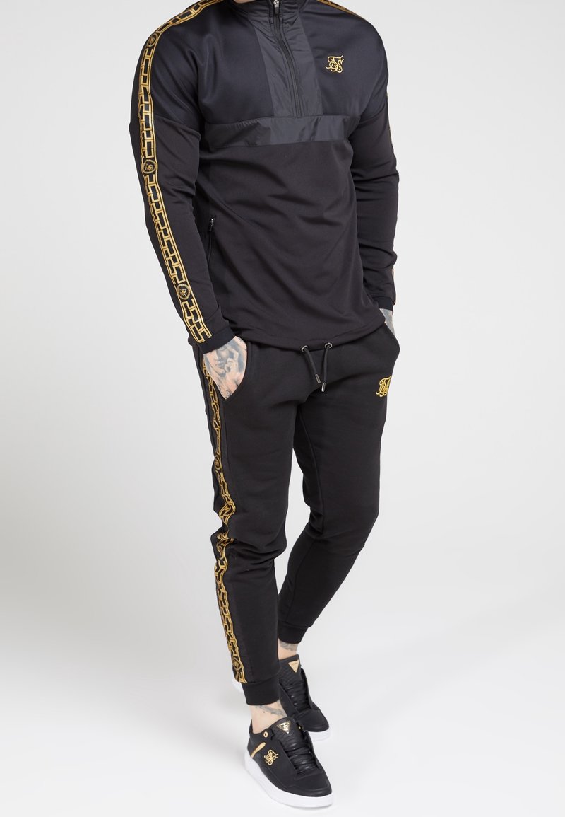 Homme en survêtement noir avec des motifs de chaînes dorées sur les manches et les côtés du pantalon, portant des baskets noires avec des accents dorés et les mains dans les poches.