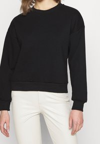 Svart crewneck-tröja med lång ärm, ribbad mudd och nederkant, tillverkad av mjukt tyg; den figurnära designen passar bra med ljusa byxor.