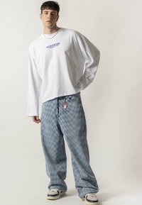 Camisa branca de manga comprida com o texto "STAY SAVAGE". Jeans azul claro xadrez oversized com um pequeno pendente de cogumelo no bolso. Ténis brancos e azul-marinho.