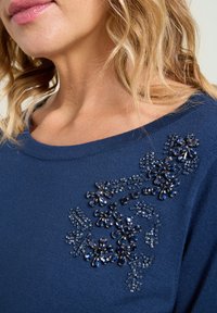 Marineblauer Pullover mit floralen Perlenverzierungen in verschiedenen Größen und Formen, aus einem strukturierten, weichen Stoff und mit weitem Ausschnitt.