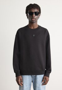 Sudadera negra con puños y dobladillo acanalados, que presenta un pequeño logo cuadrado en el pecho. Combinada con jeans de color azul claro y gafas de sol oscuras.