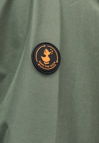 Στρογγυλό μαύρο και πορτοκαλί σήμα σε πράσινο ύφασμα που δείχνει μια καρτούν πάπια με μουσικές νότες και το κείμενο «SAVE THE DUCK», περιτριγυρισμένο από βέλη.