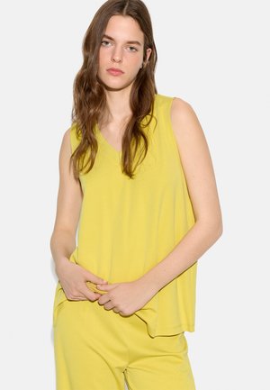 Młoda osoba z długimi brązowymi włosami, ubrana w żółty bezrękawnik i pasujące spodnie, trzymająca rant topu na jednolitym tle.