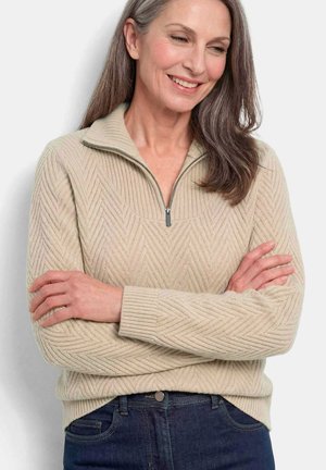 Frau mit langen grauen Haaren, die einen beigen Pullover mit Chevronmuster und Reißverschluss trägt, lächelt und verschränkt die Arme vor einem schlichten Hintergrund.