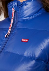 Modrá péřová bunda s vysokým límcem, vyrobená z lesklého materiálu. Obsahuje červené logo Levi's a zip s červeným a bílým táhlem.