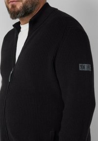 Zwarte zip-up trui met een gestructureerd rib patroon en een staande kraag, uitgerust met zijzakken en een logo accent op de linkermouw.