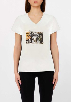 Donna che indossa una T-shirt bianca a scollo a V con collage fotografico e testo in corsivo "Walking around the world" sul davanti, abbinata a pantaloni neri.