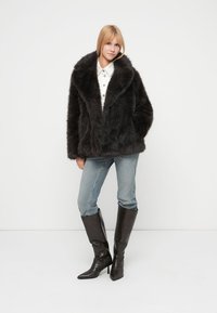 Μαύρο σακάκι από συνθετική γούνα με μεγάλο γιακά, φορεμένο πάνω από λευκό πουκάμισο και μπλε τζιν. Το μοντέλο ποζάρει με μαύρες μπότες μέχρι το γόνατο.