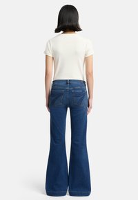 Dzwony w ciemnym denimu, z talią na poziomie średnim, tylnymi kieszeniami ozdobionymi subtelnym przeszyciem oraz gładką fakturą.