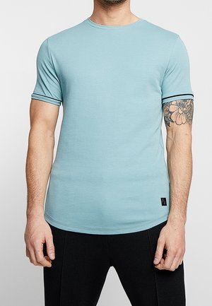 Mand iført lys blå tætsiddende kortærmet t-shirt med sort kant på ærmerne og sorte bukser, der viser en blomster-tatovering på højre arm.