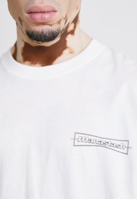 Weißes Baumwoll-T-Shirt mit Rundhalsausschnitt, versehen mit einem in Grau auf der linken Seite gedruckten "manastash"-Logo. Weiche Textur, lockere Passform.