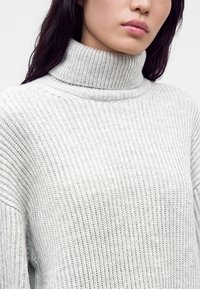 Femme portant un pull à col roulé en maille côtelée gris clair, avec de longs cheveux foncés visibles sur un fond blanc.