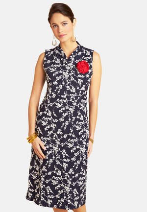 Woman wearing sleeveless navy dress with white floral pattern and large red flower brooch, accessorized with gold bracelets and hoop earrings.