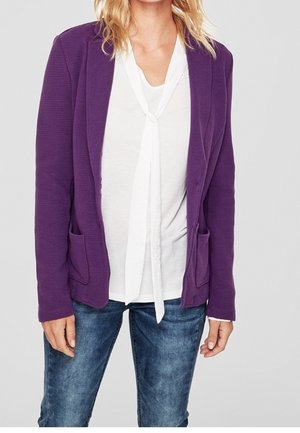 Person iført en lilla blazer over en hvid bluse med bindebånd ved halsen og blå falmede jeans, stående mod en lys grå baggrund.
