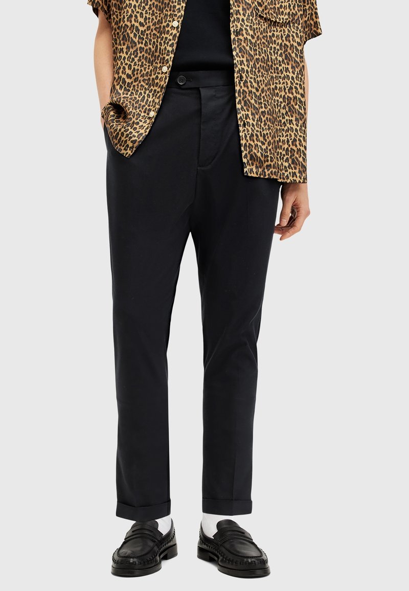 Personne portant un pantalon noir sur mesure, des mocassins noirs avec des chaussettes blanches, et une chemise à manches courtes à imprimé léopard, les mains dans les poches.