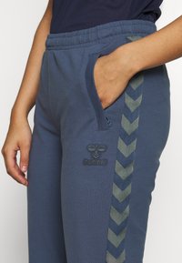 Mörkblå sweatpants i bomullsblandning med elastiskt midjeband, sidficka och gröna chevron-accenter längs sidan. Hummel-logotyp broderad.